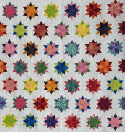 A quilt with a white background features rows of colorful, star-shaped patterns. Each star is made from various bright hues, creating a vibrant, geometric design across the entire quilt—an eye-catching piece perfect for display at a quilt show in Schenectady, NY.