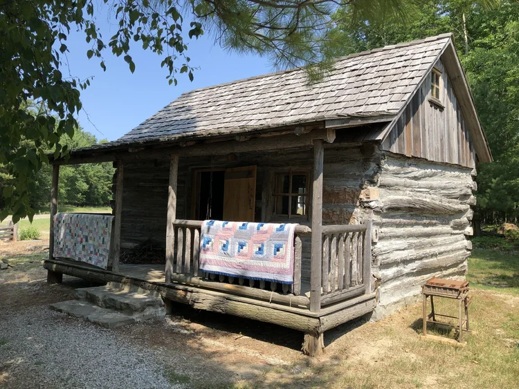 A rustic log cabin with a wooden porch, two colorful quilts draped over the railing, surrounded by trees and greenery on a sunny day—this cozy scene could easily be found during the annual quilt show in Port Austin MI.