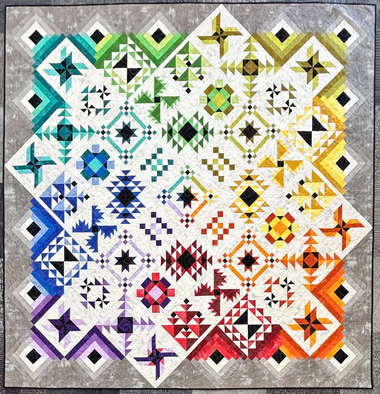 A vibrant patchwork quilt featuring geometric patterns in a gradient rainbow from blue and purple to red, orange, yellow, green, and white, bordered by a gray frame—an eye-catching piece perfect for display at a quilt show in Lafayette IN.