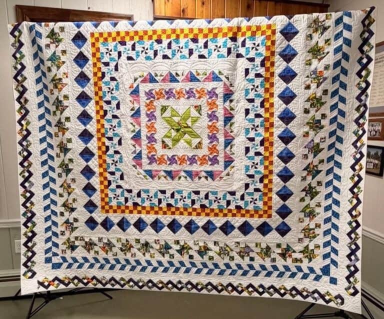A large, colorful quilt with intricate geometric patterns and multiple borders in blue, green, yellow, purple, and white is displayed on a stand against a wooden and white wall background. Discover more at https://www.esqg.com/.