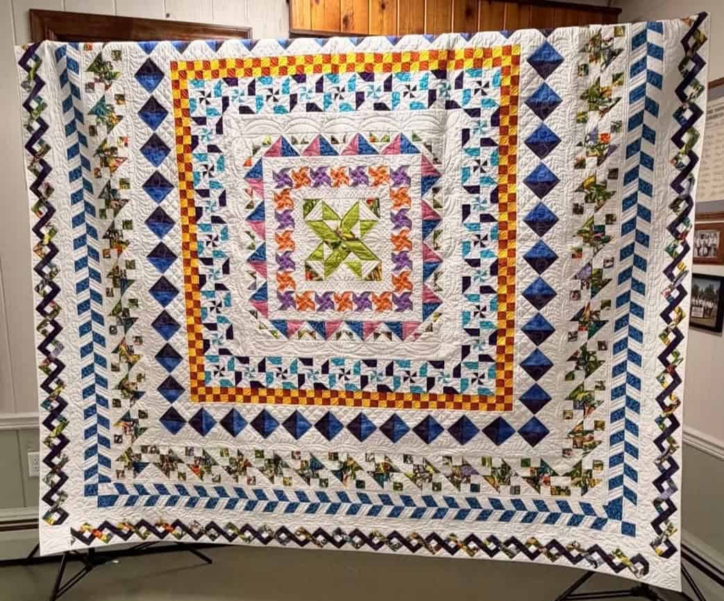 A large, colorful quilt with intricate geometric patterns and multiple borders in blue, green, yellow, purple, and white is displayed on a stand against a wooden and white wall background. Discover more at https://www.esqg.com/.