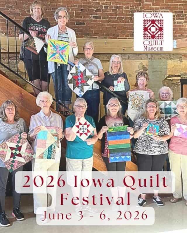 A group of women stand on stairs holding colorful quilt squares. A sign for the Iowa Quilt Museum is visible, promoting the 2026 Iowa Quilt Festival, a celebrated quilt show in Winterset IA, June 3-6, 2026.