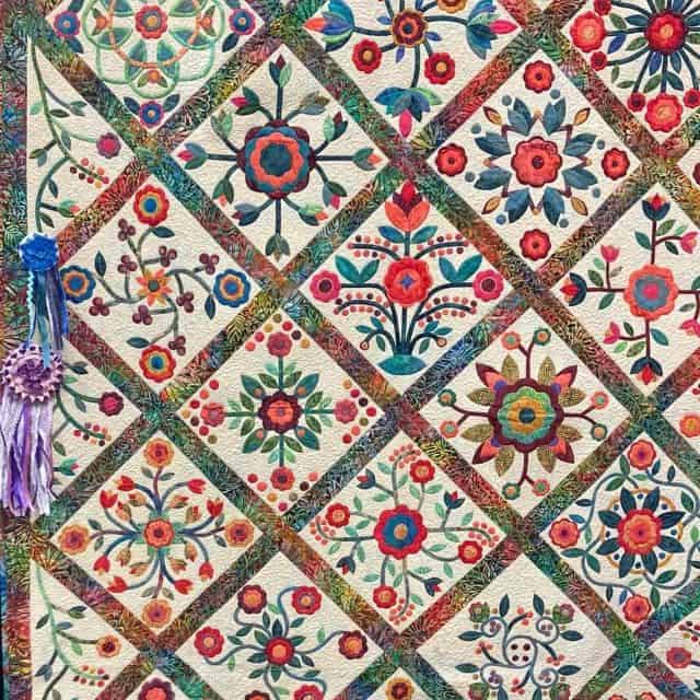 anderson-raffle-quilt A colorful quilt with a diagonal grid pattern, featuring squares filled with intricate floral and geometric designs in red, blue, green, and orange—perfect for display at a quilt show in Anderson SC. Two rosette ribbons with purple tassels hang on the left side.