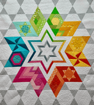 A colorful quilt, perfect for the Quilt Show in Virginia Beach VA, features a large central star made of smaller multicolored geometric patterns in blue, green, yellow, orange, red, and pink on a gray and white background.