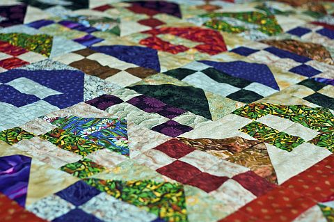 A close-up of a colorful patchwork quilt featuring various geometric shapes and patterns in shades of purple, green, red, blue, and cream—just like those displayed at the Quilt Show in Kalona IA. The quilt pieces form a vibrant, textured design.