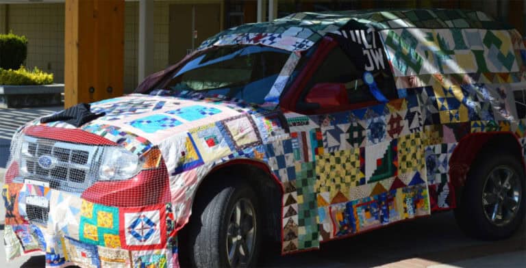 A red SUV is covered with a colorful patchwork quilt made of various fabric squares and patterns, parked outside on a sunny day during the Quilt Show in Peticon, BC, Canada.
