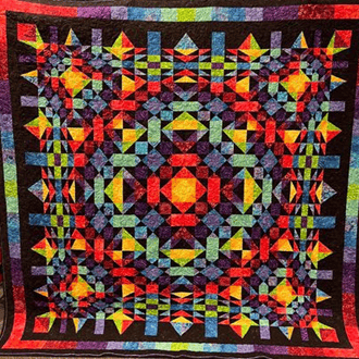 A vibrant, multicolored quilt featuring intricate geometric patterns is showcased at the Quilt Show in Enumclaw WA, with reds, oranges, greens, blues, and purples symmetrically arranged on a dark background for a striking kaleidoscopic effect.