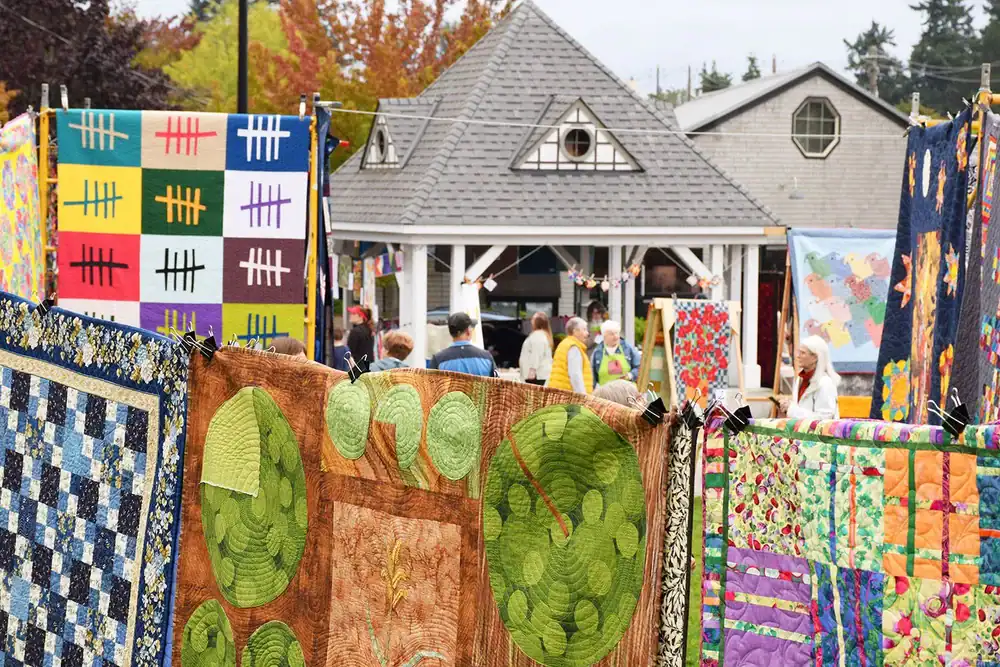 Colorful quilts hang outdoors at a lively Quilt Show in Bainbridge WA, with people walking around and a white building with a gray roof in the background. Trees with autumn leaves frame the scene.