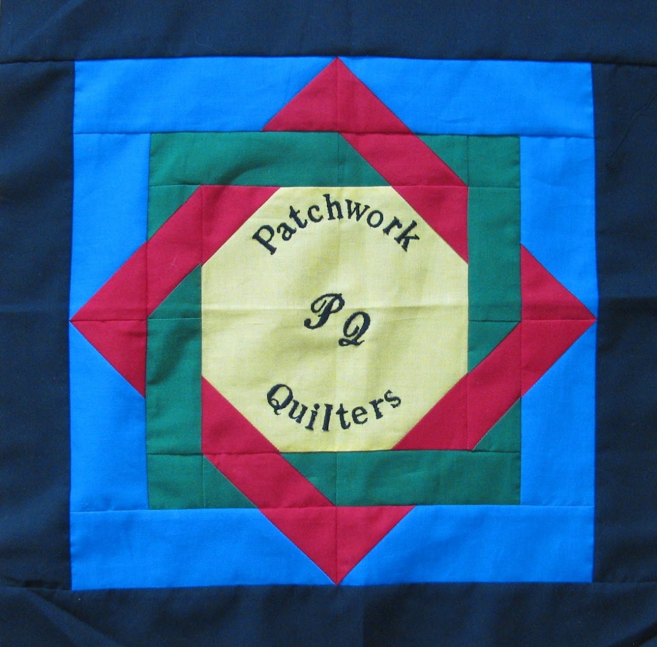 Patchwork-Quilters A colorful quilt block with concentric diamond and square shapes in blue, green, red, and yellow. The yellow center is embroidered with “Patchwork Quilters PQ.” Perfect for showcasing at the Quilt Show in Bluefield VA.