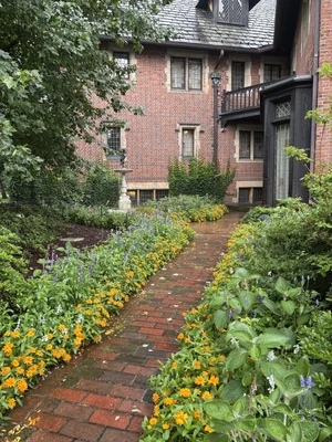 A brick pathway lined with yellow flowers and green plants leads to a red brick house with multiple windows and a small balcony, surrounded by lush foliage—an inviting scene reminiscent of the charming settings often featured at the Needlework Show in Akron OH.