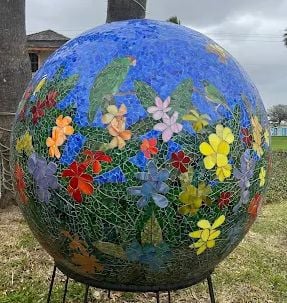 A large mosaic sphere decorated with colorful flowers and green parrots set against a blue background, displayed outdoors on a metal stand with grass and trees in the background.