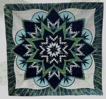 Making-Memories-Quilt-Show A quilt featuring an intricate starburst pattern in shades of blue, green, white, and black, with pointed, layered shapes radiating from the center and a leafy design along the border.