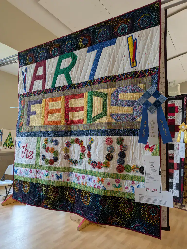 art-feed-the-soul A colorful quilt hangs on display with the words ART FEEDS the SOUL in large letters. Award ribbons, including a blue first-place ribbon, are attached to the quilt, which features various patterns and floral designs.