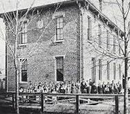 Black and white photo of a large group of children standing outside a two-story brick school building with tall windows and leafless trees in front.