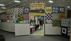 A display area with colorful quilts hanging on walls and a sign reading Quilters Club. People are inside the room, looking at more quilts and items on tables. The setting appears to be a community event or fair.