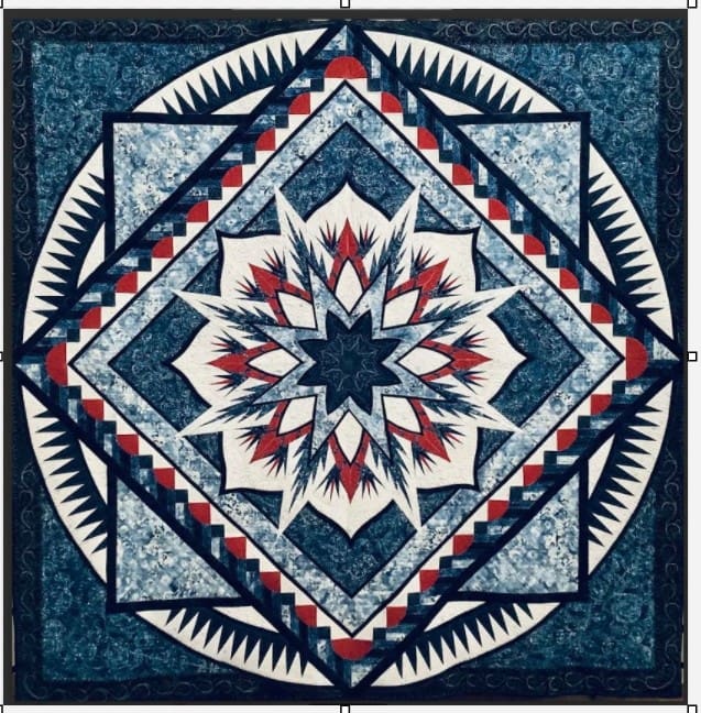 Raffle Quilt 2026 2 A patchwork quilt with geometric patterns in shades of blue, white, red, and black, featuring a large central starburst design surrounded by intricate triangles and repeating square motifs.