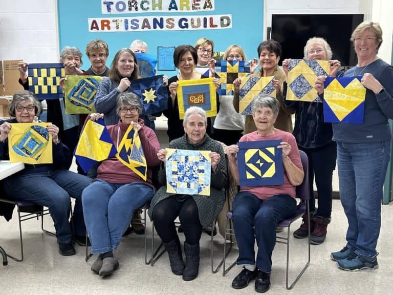 A group of women smile and display blue and yellow quilt blocks while sitting and standing in a room with a “Torch Area Artisans Guild” sign on the wall behind them.