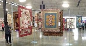 Several colorful quilts hang on display in a large, well-lit indoor exhibition hall at the Quilt Show in Port Orchard Washington. A few people admire the varied patterns and designs, with the shiny floor and curved ceiling adding to the setting.