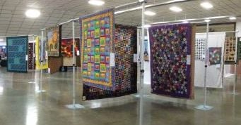 Colorful quilts hang on display racks in a spacious, well-lit room with a shiny floor, showcasing intricate designs at the Quilt Show in Port Orchard, Washington.
