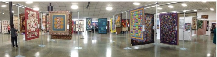 West-Sound-Quilters-Show Colorful quilts are displayed on panels throughout a large, well-lit exhibition hall with a polished floor and high ceiling. Attendees at the Quilt Show in Port Orchard Washington admire the various intricate patterns and designs.