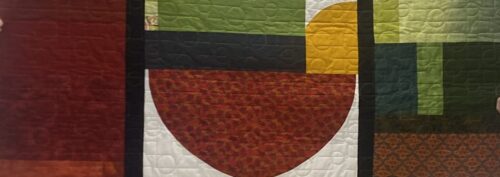 A quilt with large geometric shapes and blocks in green, red, and yellow tones, including a semicircle and square patterns, divided by thick black lines. The fabric is textured with a stitched swirling pattern.
