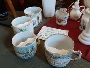 Four ornate Victorian-era moustache cups with porcelain guards are displayed on a table at the Quilt Show in Columbus, Wisconsin, accompanied by other ceramic items and a handwritten sign on a notecard.