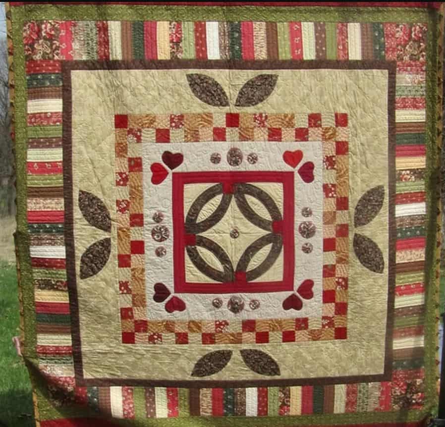 A handmade quilt with a central floral pattern, heart and leaf motifs, surrounded by red, green, and cream patchwork borders, displayed outdoors at the Quilt Show in Roxbury New York.