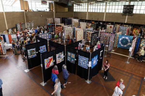 People walk among colorful quilts displayed on black partitions at an indoor Quilt Show in Amherst, Massachusetts, held in a gymnasium, with natural light coming through large windows and various groups examining the artwork.